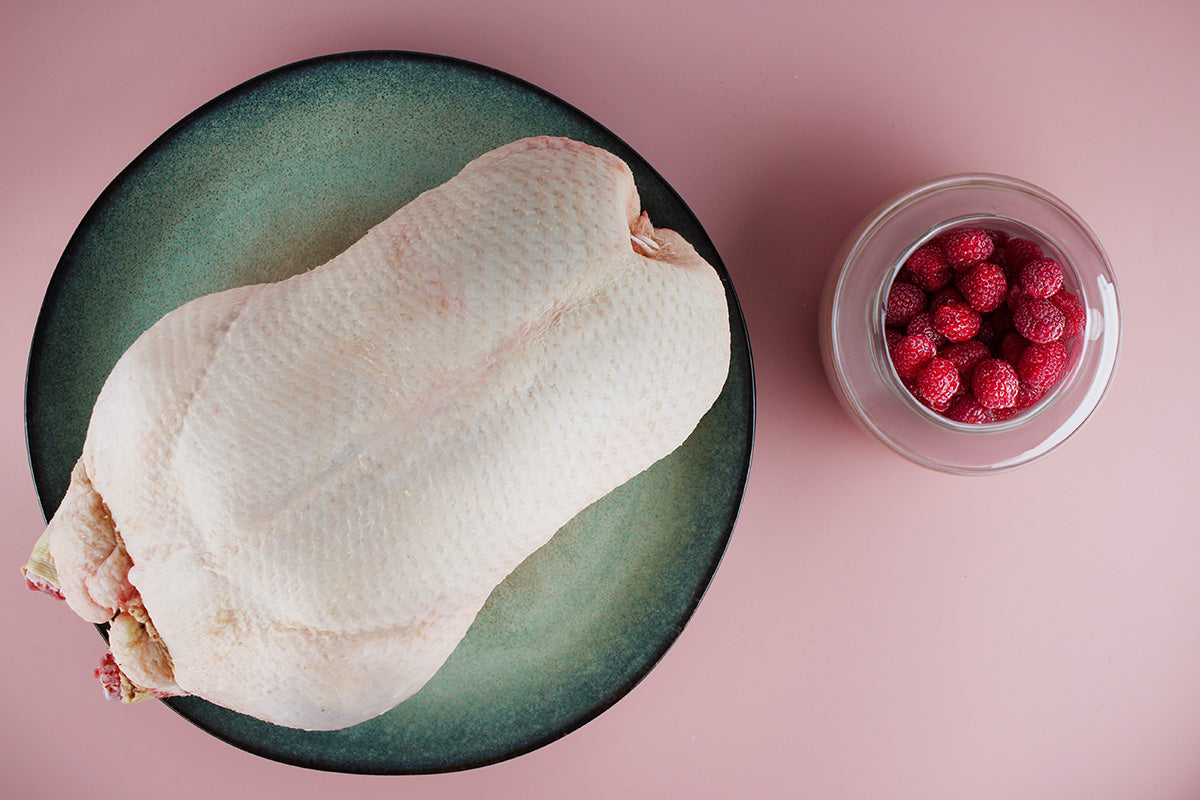 Unbehandelte Barbarieente auf grün-blauem Teller neben Glas mit Himbeeren auf rosa Untergrund.