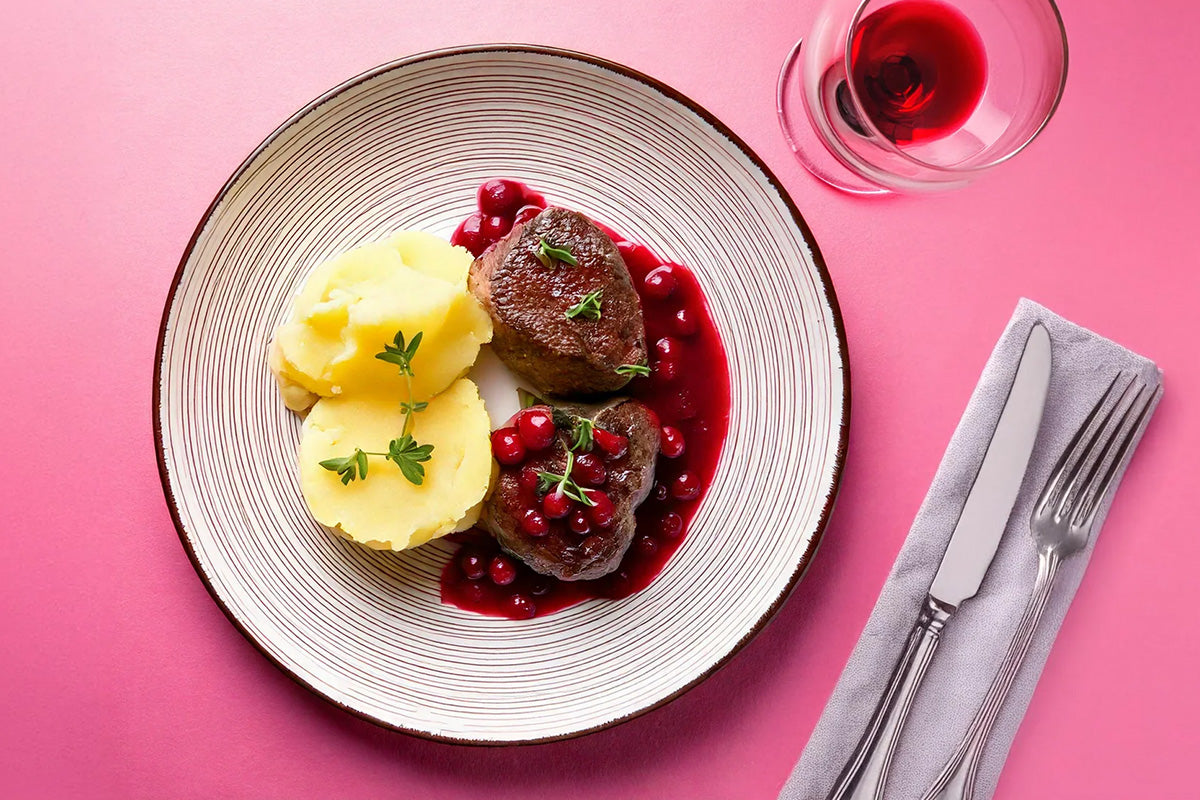Zwei Rehmedaillons mit dunkler Preiselbeersauce und Kartoffelpüree, garniert mit frischen Kräutern auf einem weißen Teller, daneben Besteck und ein Glas Rotwein auf rosa Untergrund