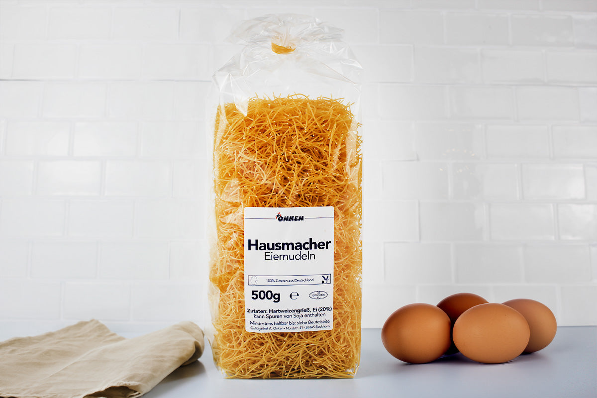 Packung mit hausgemachten Eiernudeln neben drei braunen Eiern auf weißer Oberfläche vor weißer Kachelwand