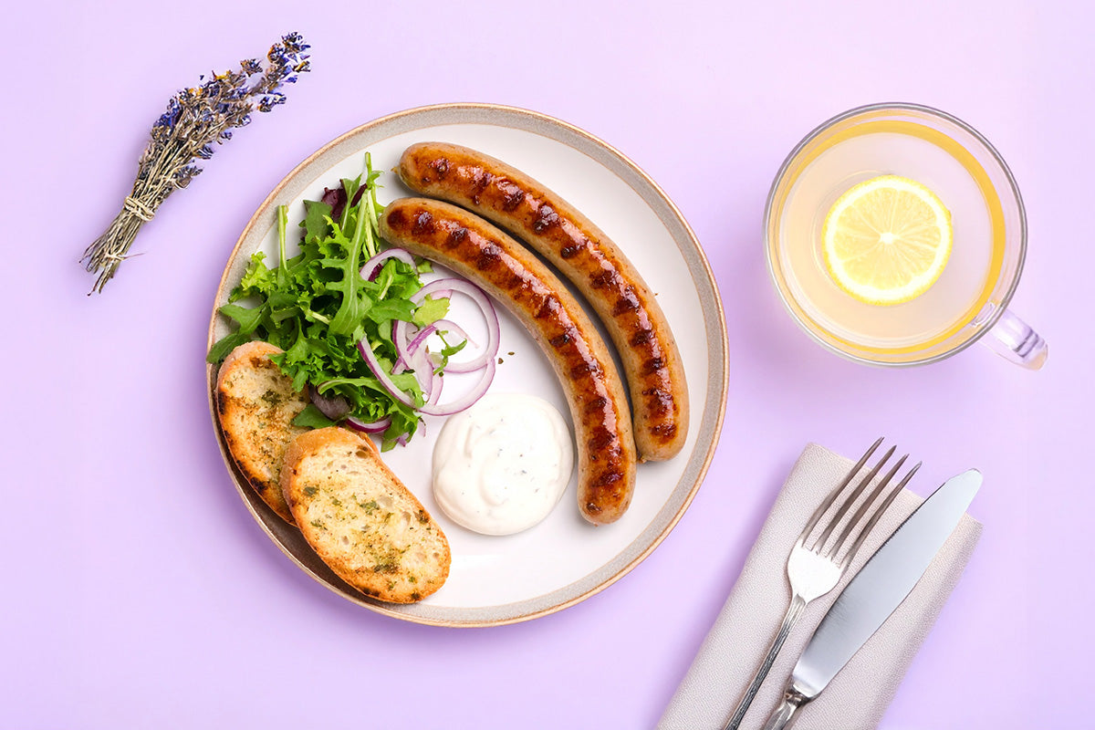 Zwei gebratene Geflügel-Chipolatawürstchen auf weissem Teller mit grünem Salat, roten Zwiebelringen, Knoblauchbrot und weißer Sauce, daneben Tasse mit Zitronenscheibe und Besteck auf hellviolettem Untergrund