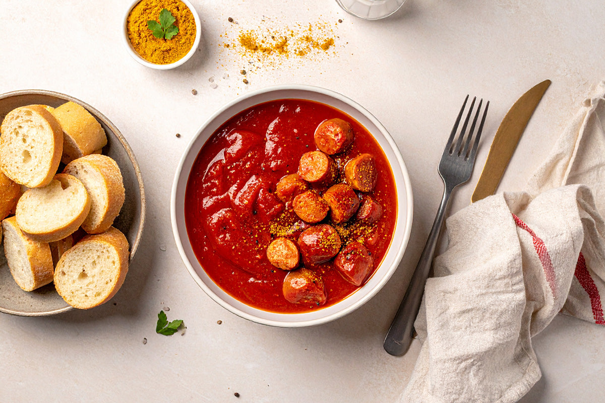 Schale mit rotem Currysoße und in Scheiben geschnittener Wurst daneben Teller mit Baguettescheiben, Besteck und Stoffserviette