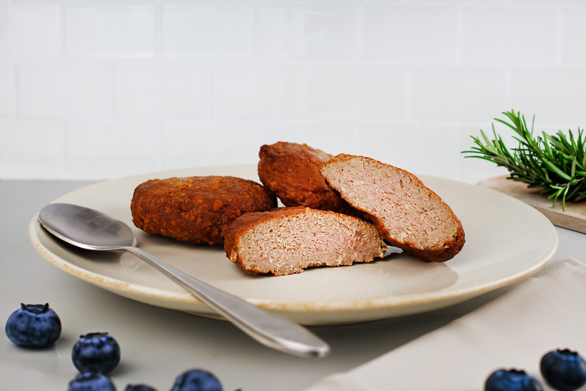 Drei Geflügelfrikadellen auf einem Teller, eine halbiert, daneben ein Löffel und einige Blaubeeren