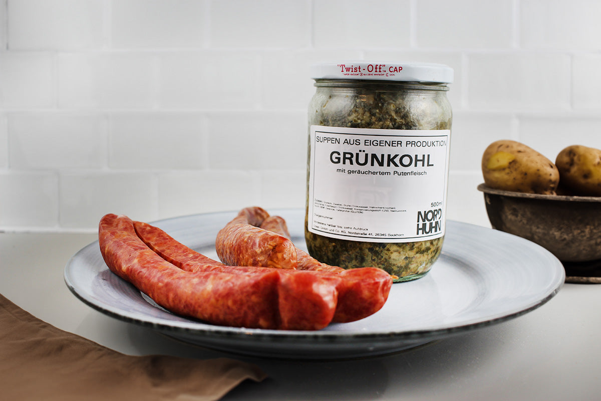 Glas mit Grünkohl und geräuchertem Putenfleisch neben zwei Würsten und zwei Kartoffeln auf einem weißen Teller