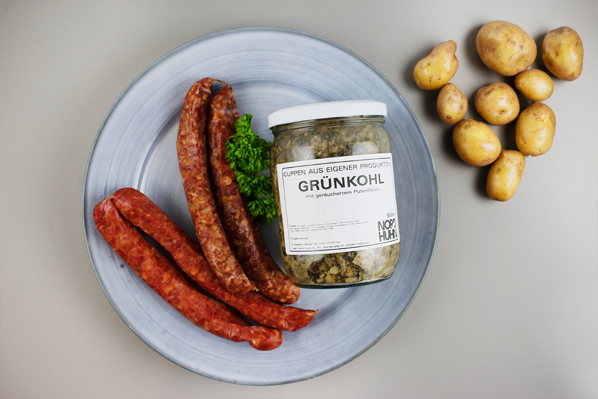 Blauer Teller mit drei Würstchen und einem Glas Grünkohl neben einer Gruppe kleiner Kartoffeln auf hellem Untergrund