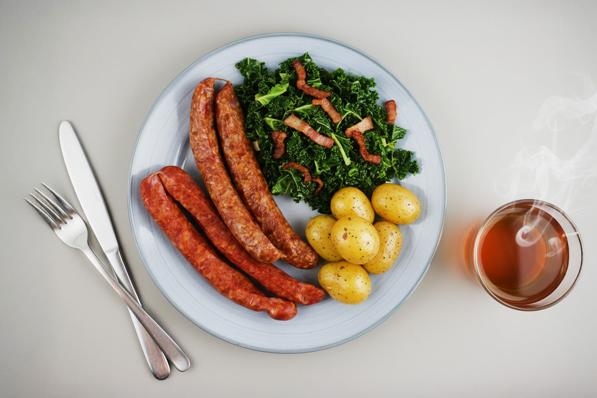 Teller mit drei Bratwürsten, Grünkohl und kleinen Kartoffeln daneben Besteck und dampfende Tasse Tee