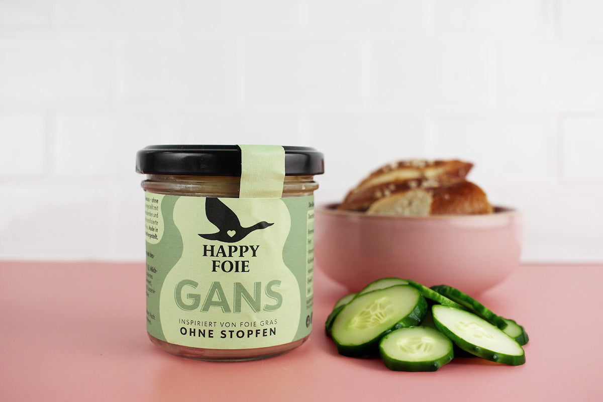 Glas Happy Foie Gans mit schwarzem Deckel, daneben Gurkenscheiben und eine Schale mit Brot auf rosa Untergrund