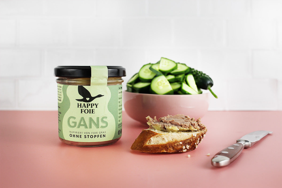 Glas Happy Foie Gans Pastete, Scheibe Brot mit Pastete, Messer und Schüssel mit Gurkenscheiben auf rosa Untergrund