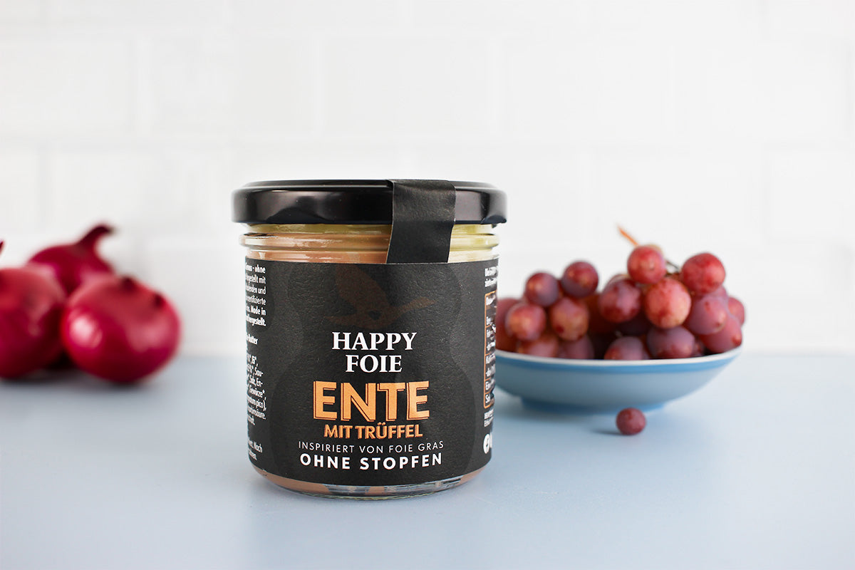 Glas mit schwarzem Etikett 'Happy Foie Ente mit Trüffel' neben rohen roten Zwiebeln und einer Schale mit roten Trauben