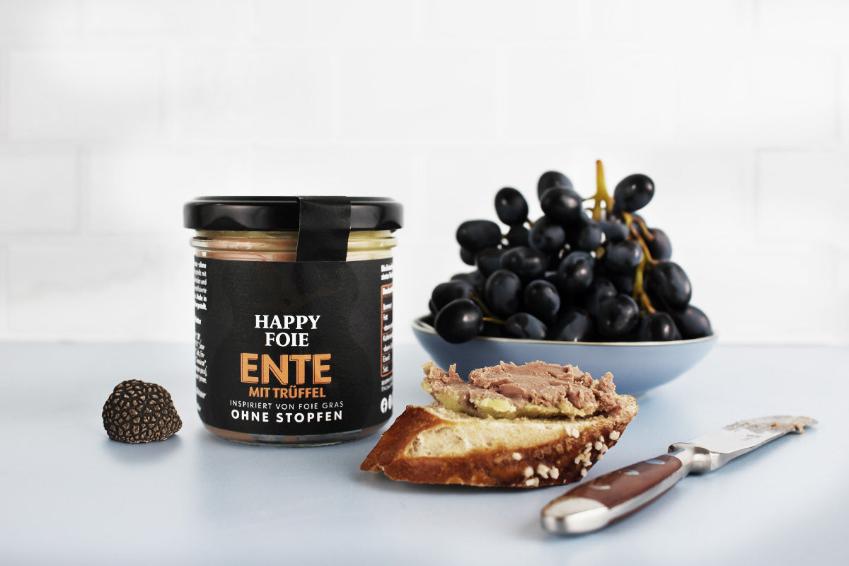 Glas Happy Foie Entenpastete mit Trüffel, daneben ein Trüffel, ein Stück Brot mit Pastete und ein Messer, im Hintergrund eine Schale mit schwarzen Trauben