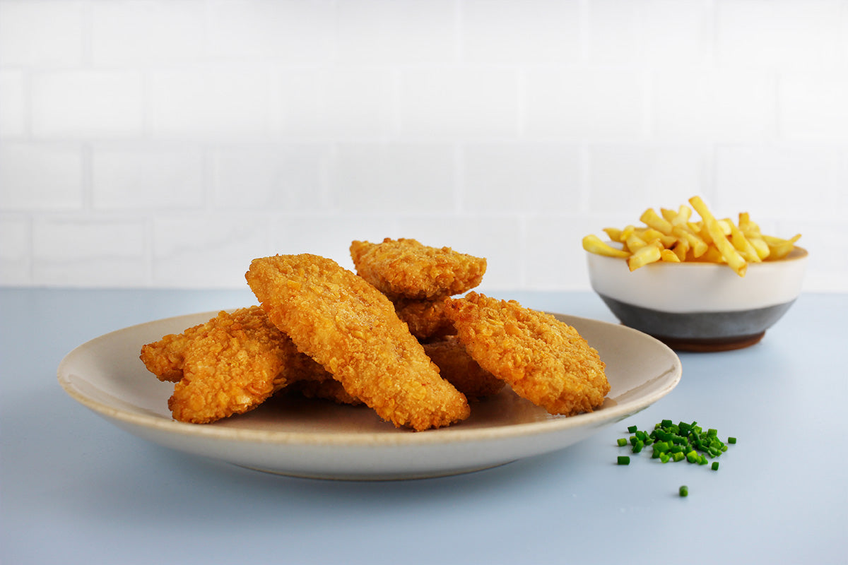 Sieben knusprige Chicken Nuggets auf einem weißen Teller, daneben eine Schale mit Pommes frites und etwas gehackter Schnittlauch auf dem Tisch