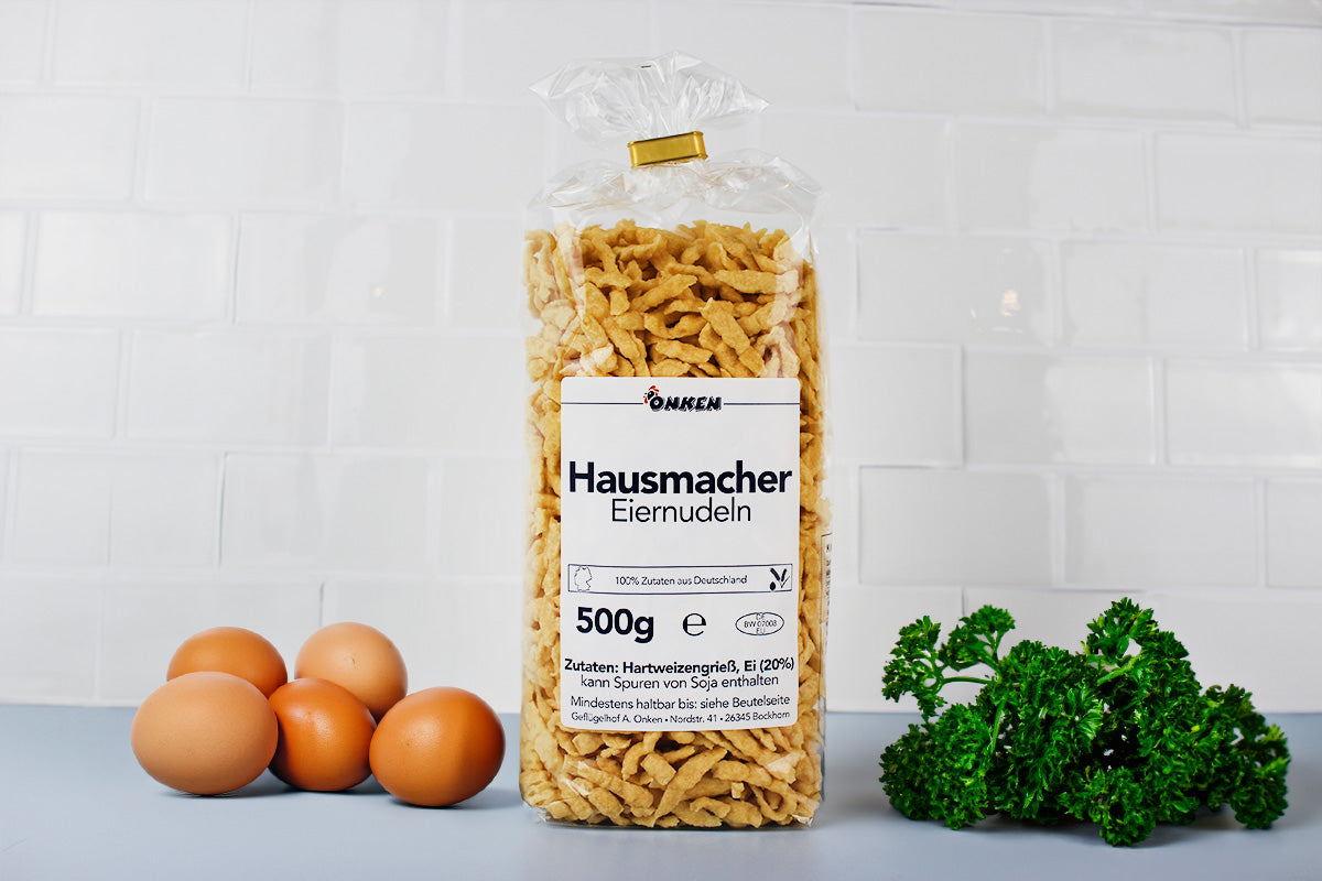 Packung mit 500g Hausmacher Eiernudeln, daneben drei braune Eier und ein Bund Petersilie auf grauer Fläche vor weißer Kachelwand