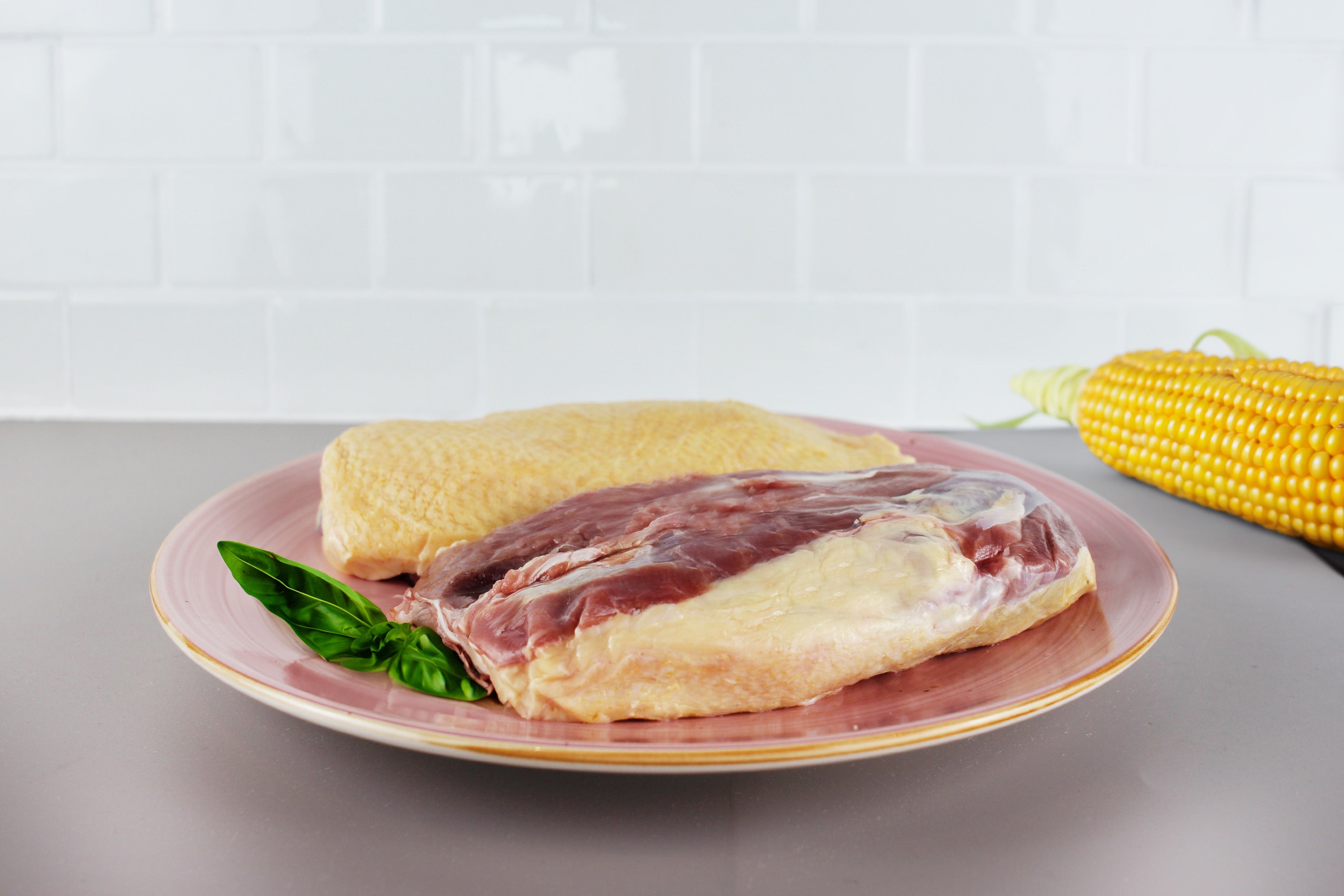 Zwei rohe Entenbrustfilets auf einem rosa Teller, daneben ein Maiskolben und ein Basilikumblatt.