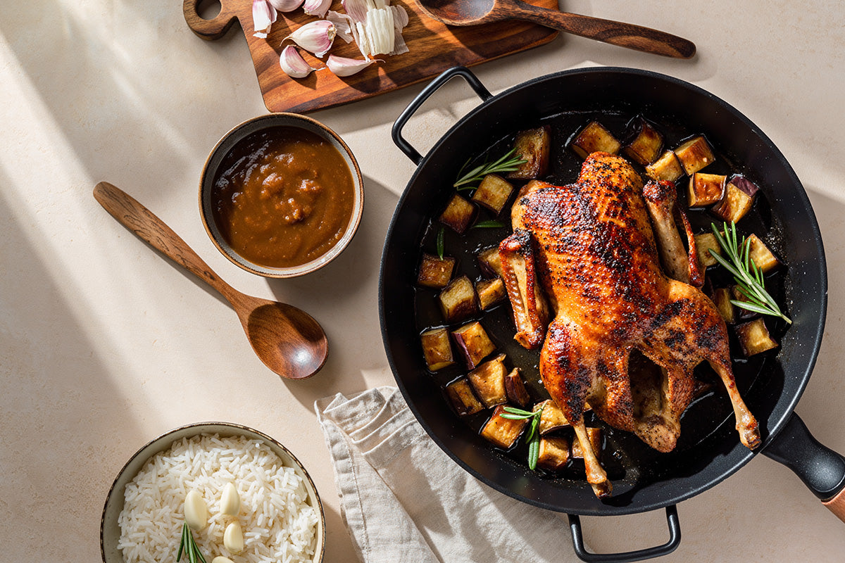 Gegrillte Ente in Pfanne mit gebratenen Kartoffelwürfeln und Rosmarinzweigen, daneben Schüssel mit Reis und Knoblauch sowie Schüssel mit brauner Sauce und Holzlöffel.