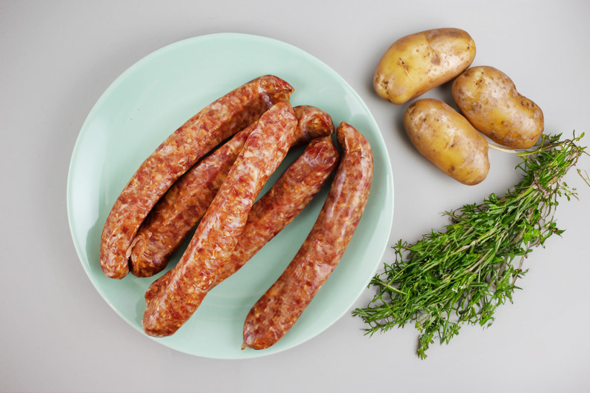 Vier rohe Würste auf einem Teller neben drei Kartoffeln und einem Bund frischer Kräuter