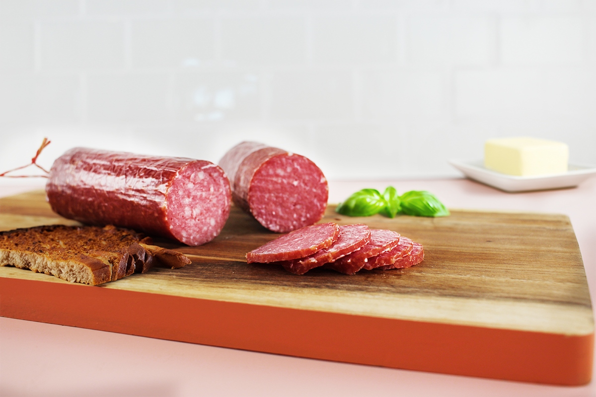 Aufgeschnittene Geflügelsalami und mehrere Scheiben auf einem Holzbrett neben zwei Scheiben Vollkornbrot und Basilikumblättern.