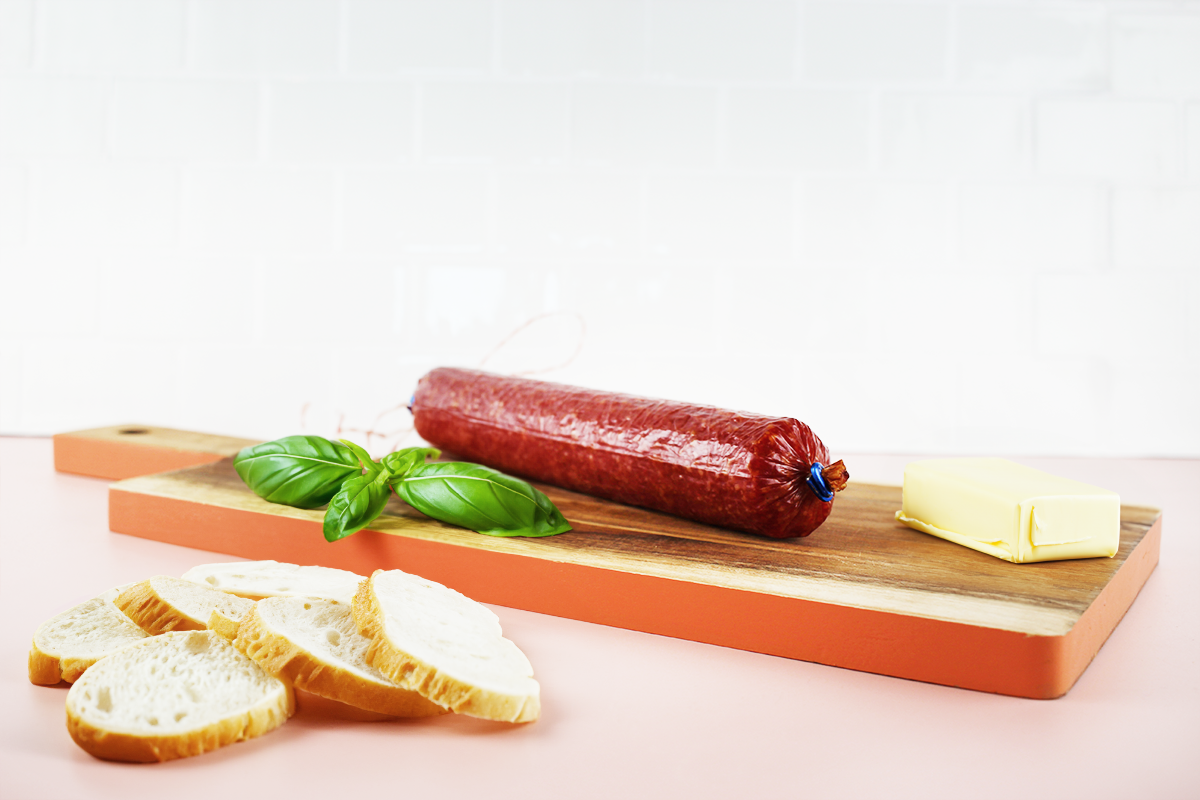 Rote Geflügelsalami auf Holzbrett mit Butterstück und Basilikumzweig, daneben mehrere Brotscheiben.