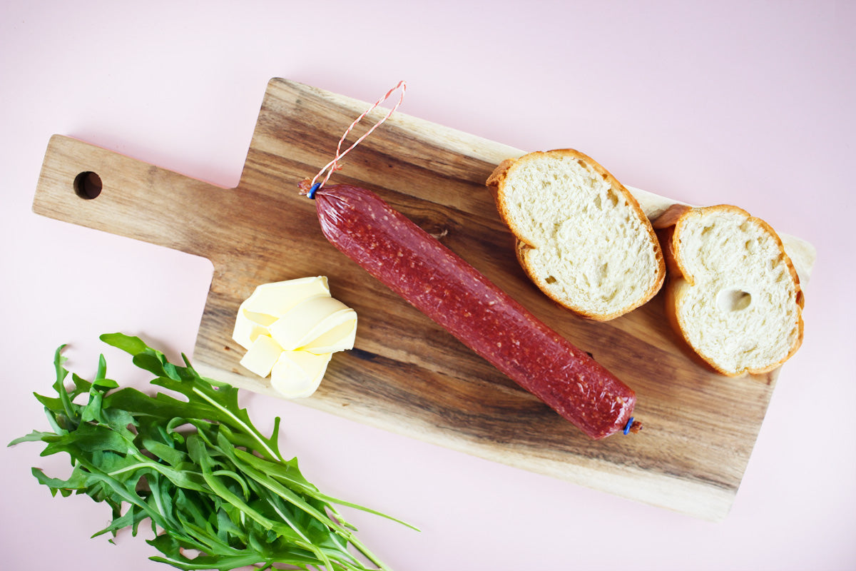 Holzbrett mit einer ganzen roten Salami, zwei Scheiben Weißbrot, Butterstücken und Rucolablättern auf hellem Untergrund.