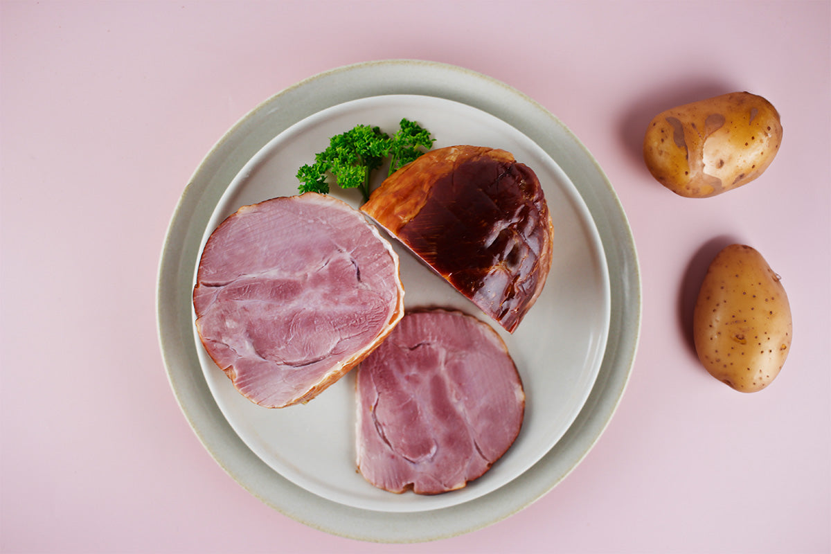 Auf einem weißen Teller liegen zwei Scheiben Putenbackschinken und ein Stück Schinken mit dunkler Kruste, daneben zwei rohe Kartoffeln und ein kleines Stück Petersilie.
