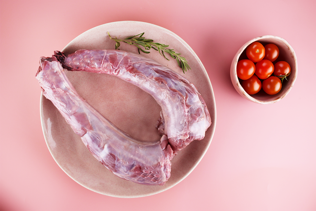 Zwei rohe Putenhälse auf einem Teller mit Rosmarinzweig, daneben Schale mit Kirschtomaten auf rosa Untergrund