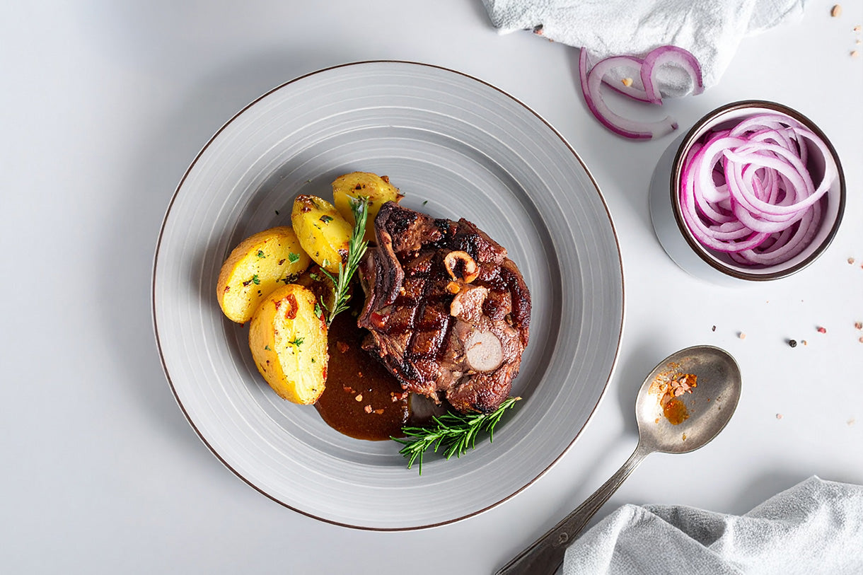 Gegrillte Rehscheibe mit Rosmarin, Bratkartoffeln und brauner Sauce auf grauem Teller, daneben rote Zwiebelringe in Schale und benutzter Löffel 