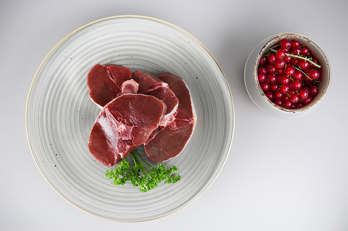Drei Scheiben rohes Rehrückenfleisch auf einem grauen Teller mit Petersilie, daneben eine Schale mit roten Johannisbeeren