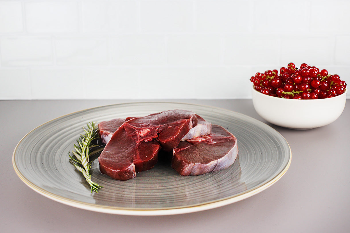 Drei rohe Scheiben Rehrückenfleisch auf grauem Teller mit Rosmarinzweig, daneben weiße Schüssel mit roten Johannisbeeren