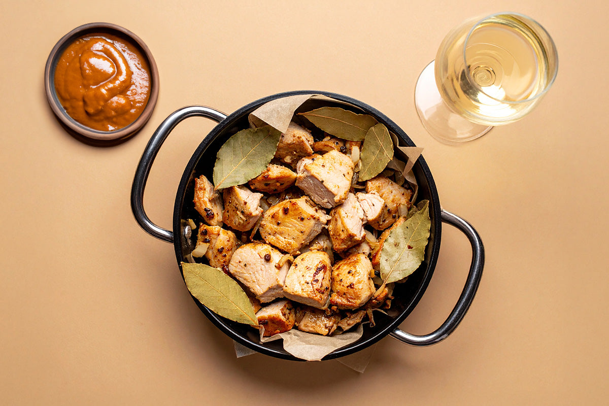 Schwarzer Topf mit gebratenen Putenfleischstücken und Lorbeerblättern, daneben Schale mit brauner Sauce und Glas Weißwein.