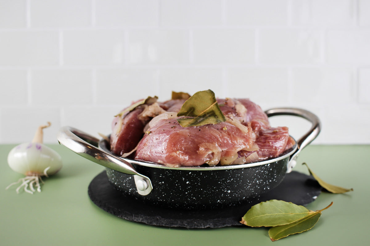 Frisches Putenfleisch mit Lorbeerblättern in einer schwarzen, marmorierten Pfanne mit Griffen, daneben eine Zwiebel und weitere Lorbeerblätter.