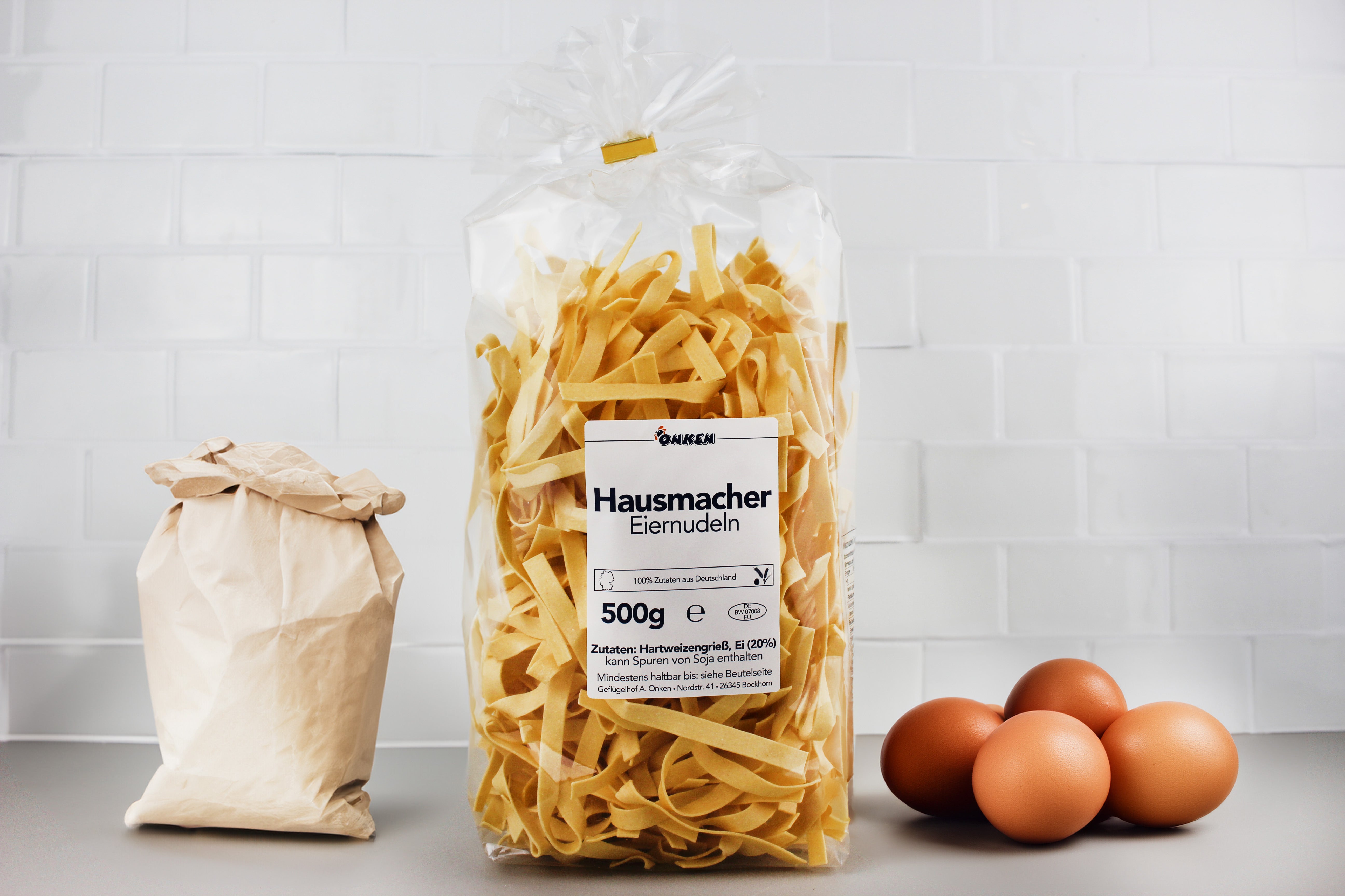 Plastiktüte mit hausgemachten Eiernudeln, Papiertüte und vier braune Eier auf grauer Oberfläche vor weißer Kachelwand