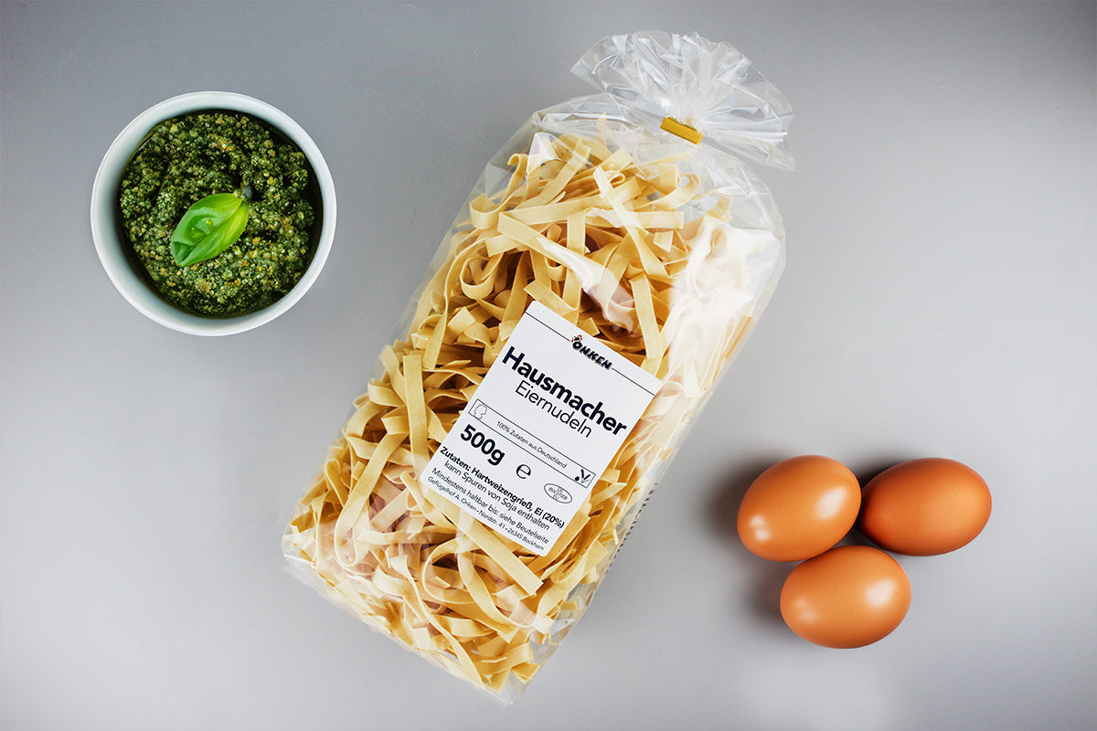 Plastiktüte mit Hausmacher Eiernudeln neben drei braunen Eiern und einer Schale mit grünem Pesto und Basilikumblatt
