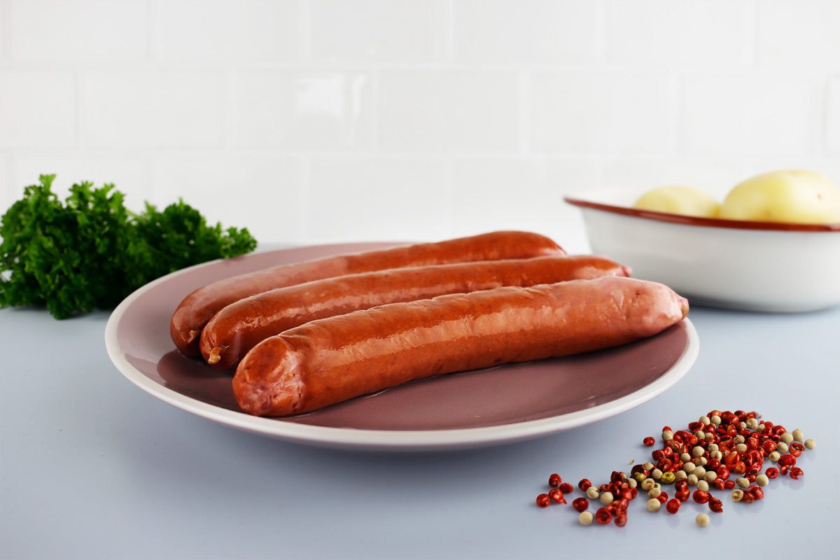 Drei lange Bratwürste auf einem Teller mit Petersilie, Zwiebeln in einer Schale und gemischten Pfefferkörnern daneben