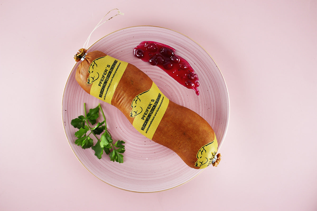 Wildschweinleberwurst mit gelbem Etikett auf rosa Teller, daneben Preiselbeeren und Petersilienzweig.